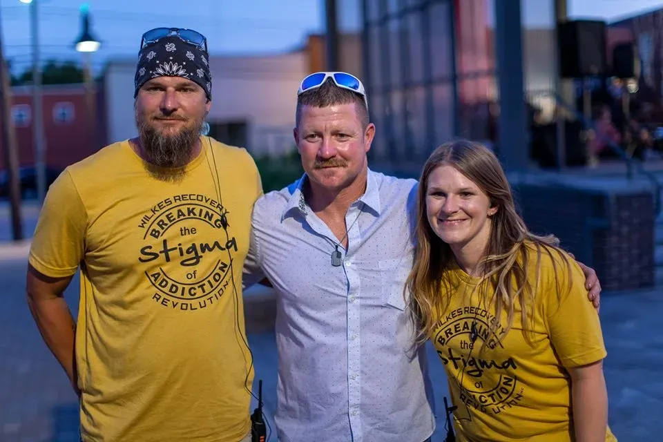 A group of friends smiles joyfully while standing in a circle outdoors, surrounded by vibrant fall foliage and soft sunlight